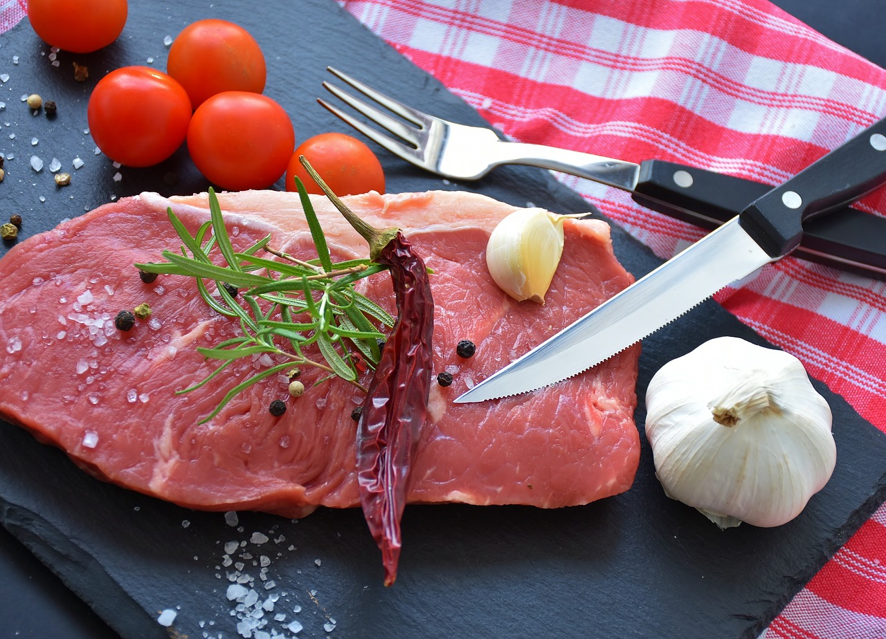 Raw steak on a black slate board with rosemary, dried chili, garlic clove, coarse salt, peppercorns, cherry tomatoes, and a knife and fork on a red checkered cloth.