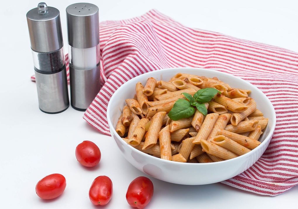 White bowl of penne pasta with tomato sauce and basil leaf on a red striped cloth, with cherry tomatoes and salt and pepper shakers nearby.